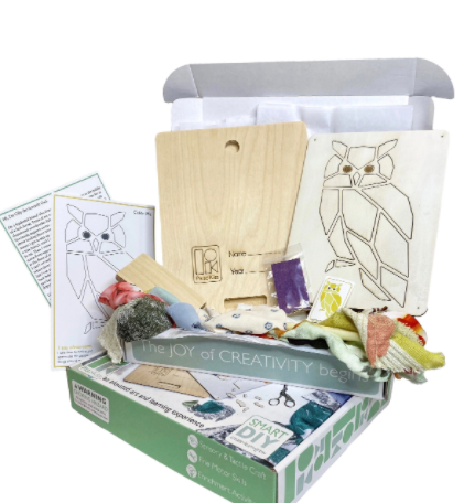 An open box contains a wooden cut-out owl and fabric scraps to be used for the craft