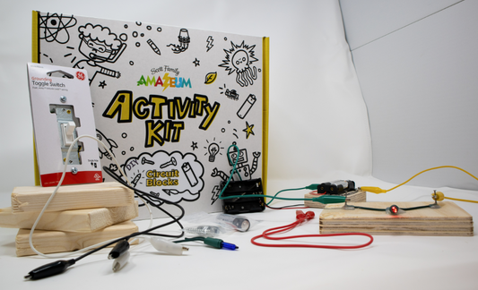 A white box with black marker-like drawings printed on the front says Scott Family Amazeum Activity Kit: Circuit Blocks. In front of the box, a circuit has been assembled with different wires, connections, switches, and batteries that are included in the kit. 